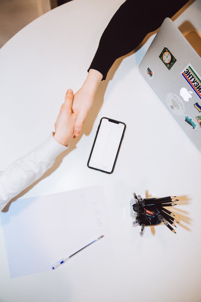 Services Two individuals shaking hands across a desk with a smartphone and laptop in a modern office setting.